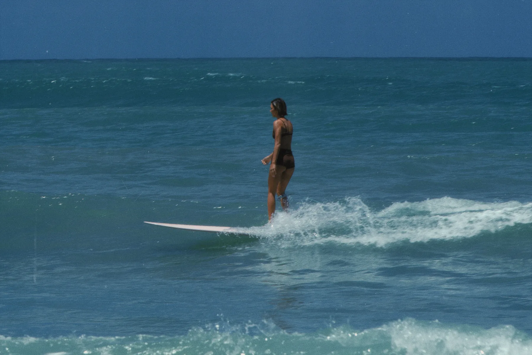 Surfer riding a longboard on a small wave under a clear sky