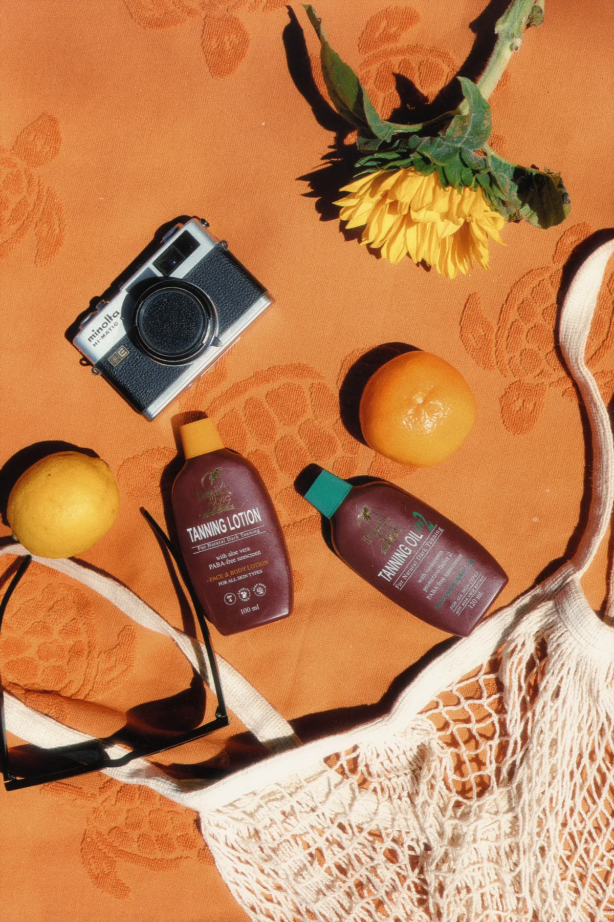 Hawaiian Style Tanning Lotion and Classic Tanning Oil on an orange beach towel with citrus fruit, a camera and a sunflower