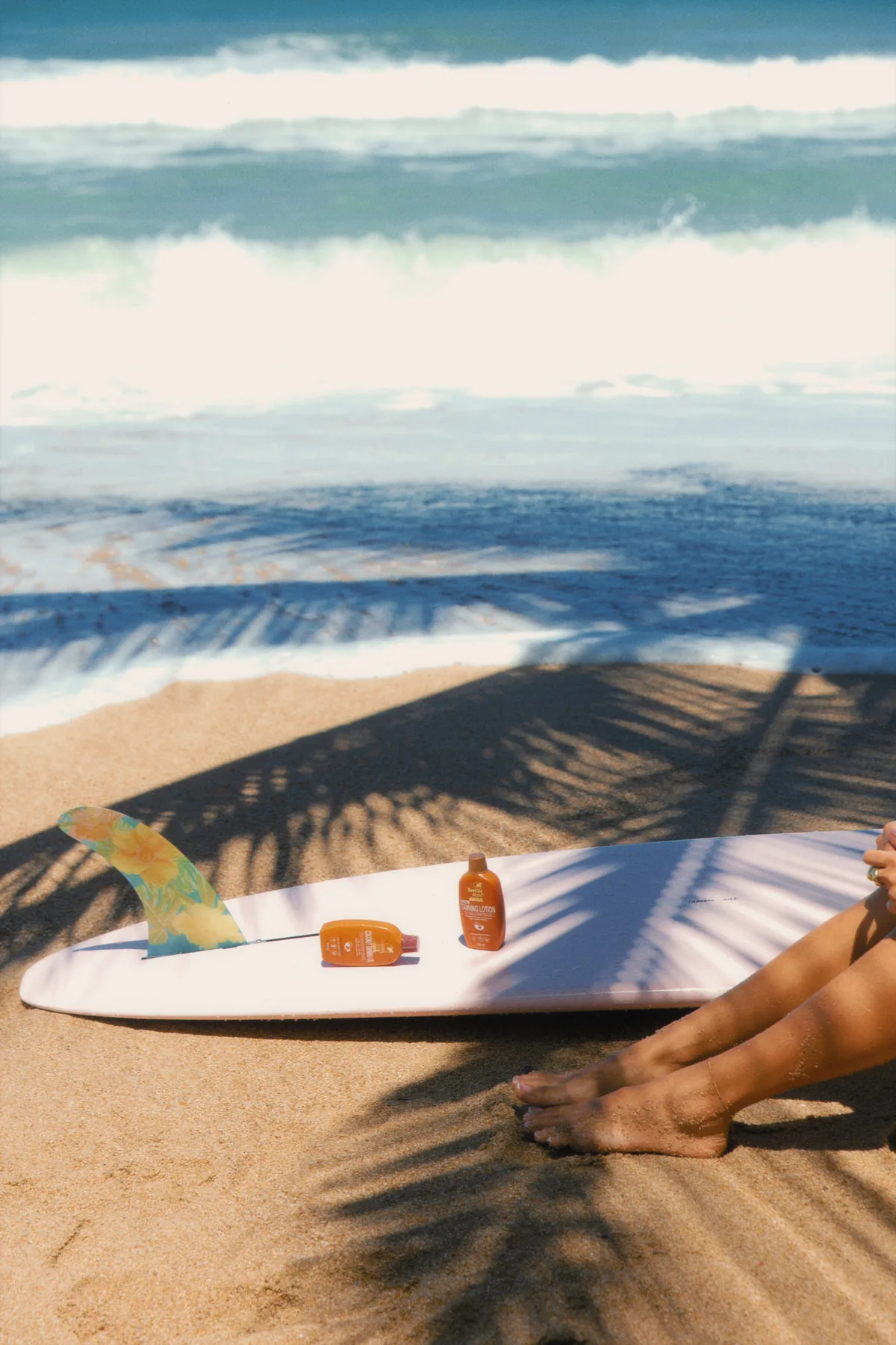 Coconut Tanning Oil and Tanning Lotion laid out on a surfboard in palm shade by the sea